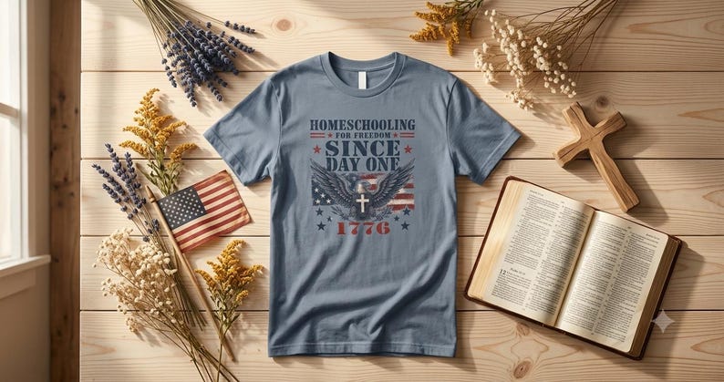 May include: A slate blue short-sleeve t-shirt featuring a graphic print that reads "HOMESCHOOLING FOR FREEDOM SINCE DAY ONE 1776" with an eagle, a cross, and American flag elements. The shirt is displayed flat with a small flag and an open book.