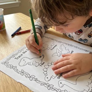 May include: A child is using a green crayon to trace a dinosaur outline on a white paper. The paper has the text "Trace the D" and features illustrations of various dinosaurs, including a T-Rex and Stegosaurus. Crayons are visible on the table.