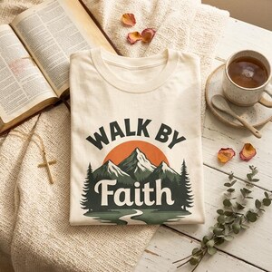 May include: A cream-colored t-shirt featuring a graphic print of a mountain range, pine trees, and a setting sun. The text "WALK BY FAITH" is printed in dark green lettering above and below the mountain illustration on the front of the shirt.