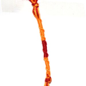 Może przedstawiać: Bransoletka z czerwonej i pomarańczowej plecionej sznurka. Bransoletka jest zawiązana na górze węzłem.