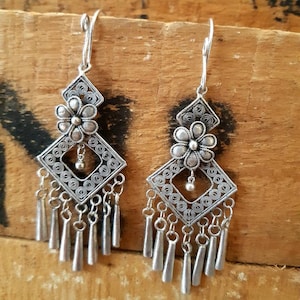 以下が含まれることがあります： 正方形のデザインと花のチャームが付いたシルバーのドロップイヤリング。イヤリングは繊細で複雑なデザインで、どんな服装にもエレガントさを加えるのに最適です。