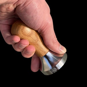 以下が含まれることがあります： 明るい茶色の木製ハンドルとステンレス鋼のベースを備えたコーヒータンパー。ベースには「53.4」の数字が刻印されています。タンパーはコーヒー粉をプレスするように設計されています。