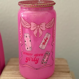 May include: A pink, glittery glass can-shaped tumbler with a decorative lid and straw. The tumbler features a pink bow and banner design with three energy drink can illustrations. The text "energy drink girly" is also visible.