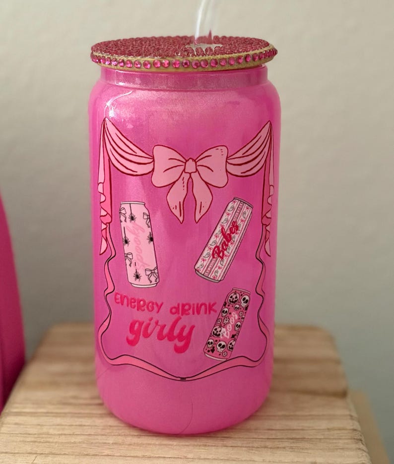 May include: A pink, glittery glass can-shaped tumbler with a decorative lid and straw. The tumbler features a pink bow and banner design with three energy drink can illustrations. The text "energy drink girly" is also visible.