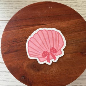 May include: A pink seashell sticker with a bow. The sticker has a glittery effect and is set against a wooden background. The seashell design features vertical lines and a bow with heart details.