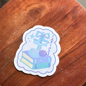 May include: A fantasy-themed sticker featuring a book with roses, a sword, and a bow. The sticker has a light blue and purple color scheme with the word "FANTASY" written vertically on the right side. The sticker is on a wooden surface.