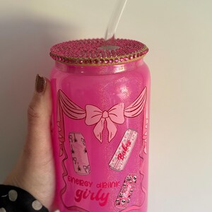 May include: A pink glass tumbler with a decorative lid and a clear straw. The tumbler features a bow and energy drink graphics with the words "Energy drink girly". The lid is embellished with pink rhinestones.