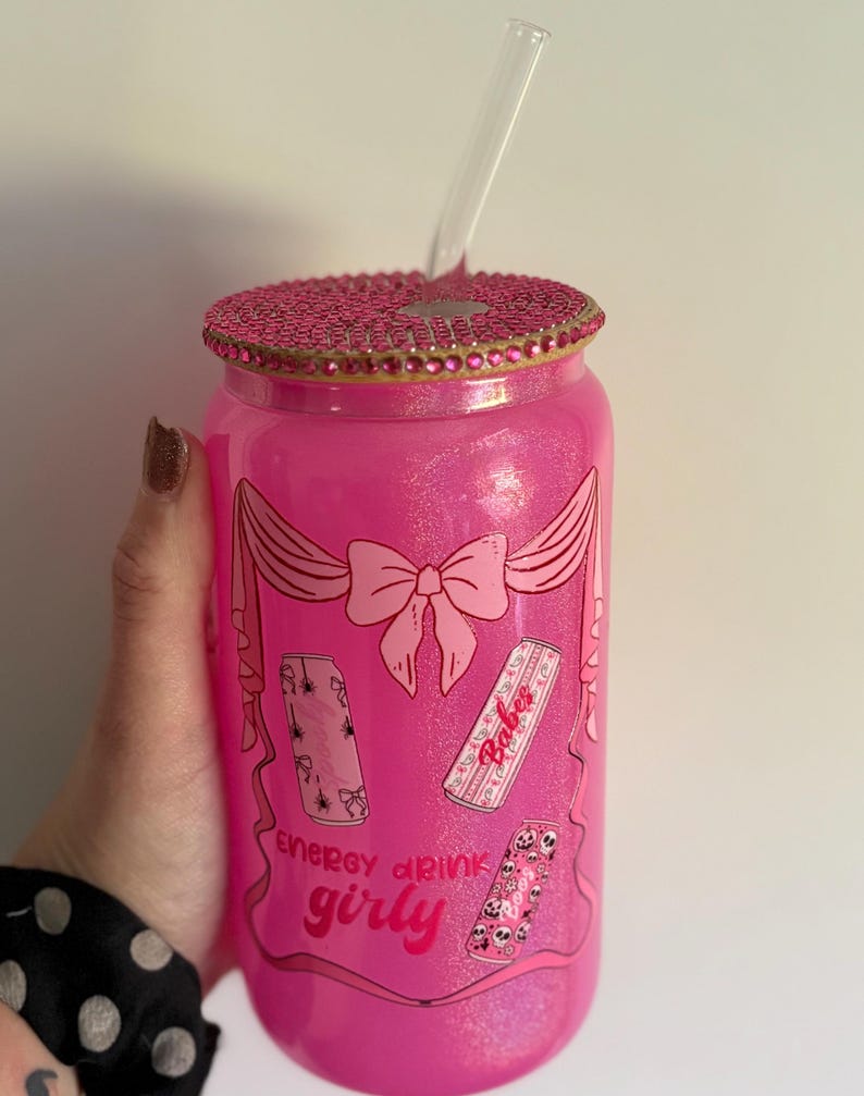 May include: A pink glass tumbler with a decorative lid and a clear straw. The tumbler features a bow and energy drink graphics with the words "Energy drink girly". The lid is embellished with pink rhinestones.