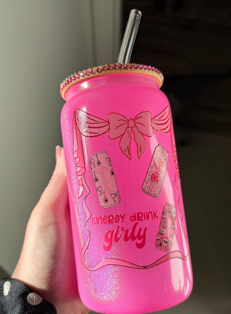 May include: Pink glittery glass can with a clear straw and a bedazzled rim. The can features a bow design and the words "energy drink girly" with illustrations of cans with spooky designs.