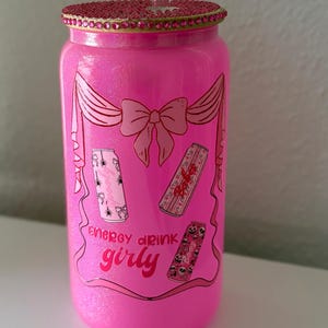 May include: A pink glittery glass can-shaped tumbler with a decorative lid and straw. The tumbler features a bow design and the words "energy drink girly" with illustrations of energy drink cans.