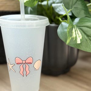May include: A clear, glitter-covered plastic tumbler with a lid and straw. The cup is decorated with a pink bow, a starfish, and a seashell. The cup is next to a green plant in a black pot.