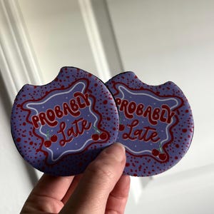 May include: Two car coasters with a purple background and the words "Probably Late" in red script. The coasters have a cherry design and a black border. The coasters are held by a hand.