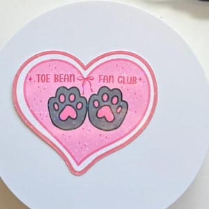 May include: A pink heart-shaped sticker with the text "TOE BEAN FAN CLUB" and two gray paw prints with pink toe beans. The sticker has a glittery effect and is on a white background.