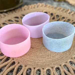 May include: Three glittery, cylindrical drink sleeves in pastel colors: pink, lavender, and blue. The sleeves are arranged on a textured, round, brown placemat. The sleeves are designed to fit around a standard-sized beverage container.
