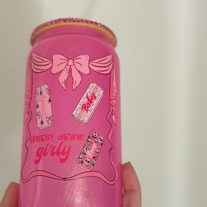 May include: A pink energy drink can with a glittery finish and a decorative rim. The can features a bow design and the words "energy drink girly" along with illustrations of smaller cans. The overall design is feminine and playful.