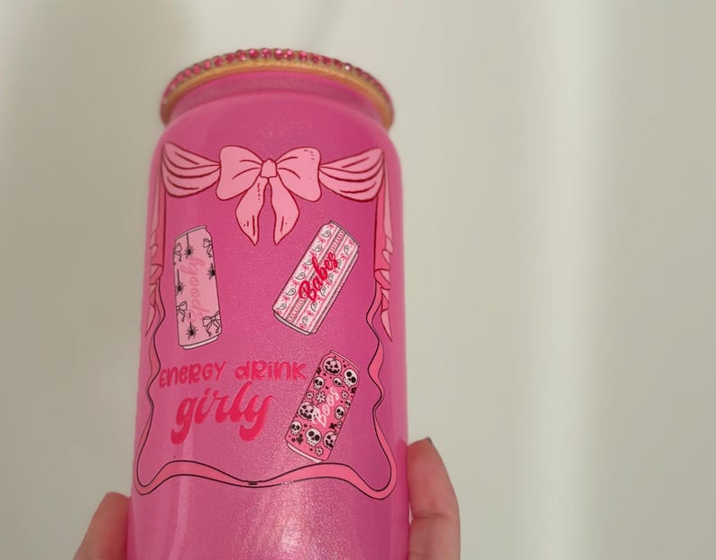 May include: A pink energy drink can with a glittery finish and a decorative rim. The can features a bow design and the words "energy drink girly" along with illustrations of smaller cans. The overall design is feminine and playful.
