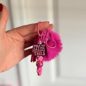 May include: A pink keychain with a fluffy pom-pom, a metal clasp, and a charm that reads "HOLD ON LET ME OVERTHINK THIS." The charm has a pink tassel and a beaded accent.