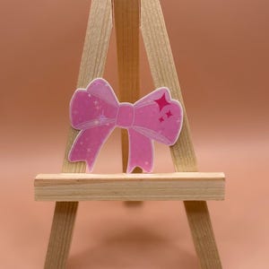 May include: A pink sparkly bow with a white background. The bow is on a small wooden easel.