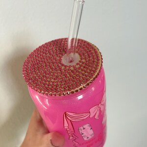 May include: A pink tumbler with a bedazzled lid and a clear straw. The lid is covered in small, pink rhinestones. The tumbler has a design with a bow and text. The tumbler is held by a hand.