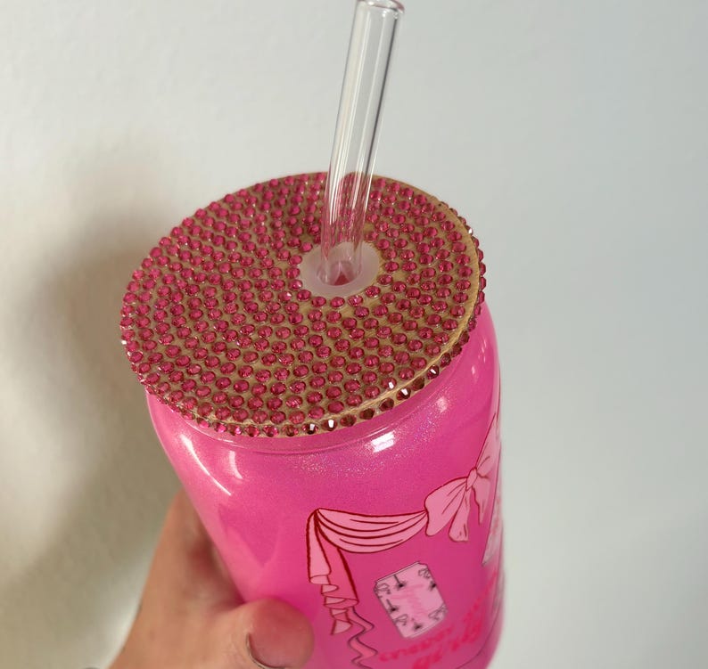 May include: A pink tumbler with a bedazzled lid and a clear straw. The lid is covered in small, pink rhinestones. The tumbler has a design with a bow and text. The tumbler is held by a hand.