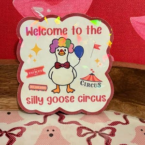 May include: A sticker with a whimsical design featuring a goose wearing a bow tie and a rainbow-colored wig, holding a blue balloon. The text reads "Welcome to the silly goose circus."