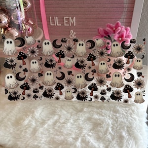 May include: A clear rectangular sheet featuring a repeating pattern of white ghosts, brown mushrooms, black crescent moons, and dark flowers. The design is printed in a grid-like arrangement across the transparent surface, showcasing a variety of spooky and botanical elements in a consistent, repeating graphic style.