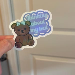 May include: A sticker featuring a brown teddy bear with a blue bow, and the text "Thinking about my bed" in a blue cloud-shaped design. The sticker has a holographic effect.