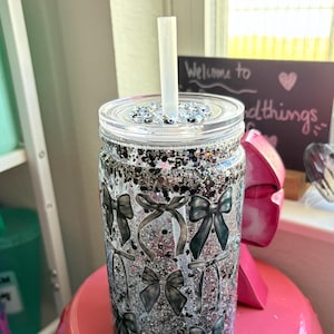 May include: A clear tumbler with a lid and straw, decorated with black and silver glitter and black bow designs. The tumbler is sitting on a pink surface. The text "Welcome to good things" is visible in the background.
