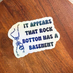 May include: A holographic sticker with the text "IT APPEARS THAT ROCK BOTTOM HAS A BASEMENT" in blue and green. A skeleton illustration is on the left side of the sticker. The sticker is on a wooden surface.