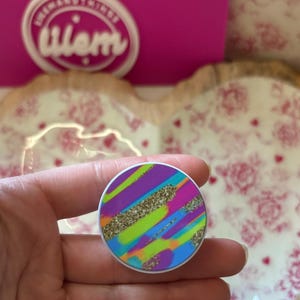 May include: A round phone grip with a colorful abstract design. The design features diagonal stripes in purple, green, blue, and orange, with gold glitter accents. The grip has a white border and is held in a hand.