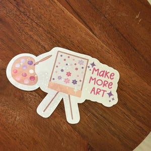 May include: A sticker featuring a painter's palette and easel with the text "Make More Art." The palette has pink and purple paint splotches. The easel has a painting with pink and purple flowers.