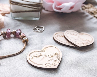 May include: A gold-toned bracelet with pink and clear beads and charms, and two wooden heart-shaped wedding favors. The favors are engraved with names and a date, and surrounded by floral designs. A pair of rings and other decorative items are in the background.