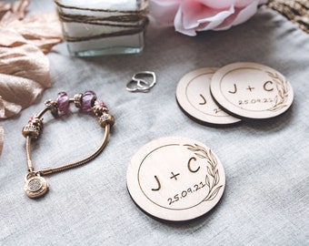 May include: Two wooden round ornaments with the text "J + C 25.09.21" and a leafy design. The ornaments are on a light grey fabric surface.