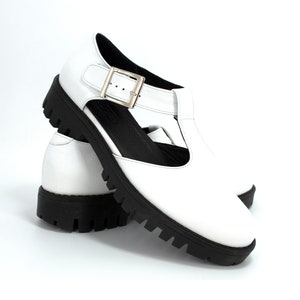 Può includere: Sandali bianchi in pelle con cinturino con fibbia e suola spessa in gomma nera.