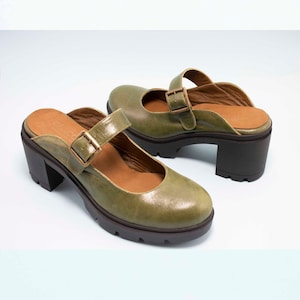 Pode incluir: Um par de mules de couro verde com salto alto e fivela. Os sapatos têm um salto plataforma grosso e uma ponta arredondada.