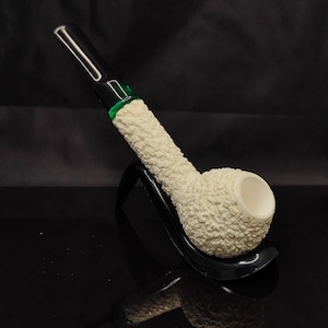 May include: A tobacco pipe featuring a textured, off-white bowl and shank with a rough, granular surface. The pipe includes a smooth, black acrylic stem connected to the shank by a translucent green band, resting on a black curved stand.