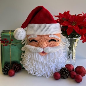 Pode incluir: Cabeça de Pai Natal de crochê com gorro vermelho, barba branca e bochechas rosadas. A cabeça do Pai Natal faz parte de uma decoração de Natal. Um presente verde embrulhado, enfeites vermelhos e um vaso de flores vermelhas estão no fundo.
