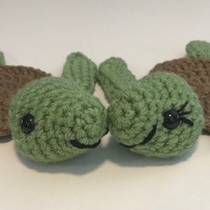May include: Two crocheted green turtle toys with brown shells and black eyes. The turtles are facing each other and smiling.