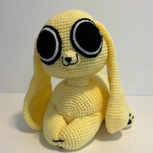May include: A crocheted yellow bunny with large black eyes and white accents. The bunny is sitting with its long ears flopped down.