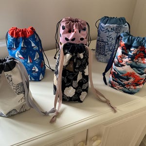 May include: Six fabric drawstring bags featuring various patterns including sailboats on blue, a crane on white, black with white flowers, pink with black shapes, blue with botanical prints, and a landscape with red trees and a mountain peak.