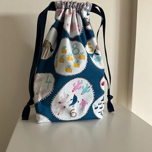 May include: A drawstring bag featuring a teal background with white circular patterns containing illustrations of animals, insects, and numbers. The bag has dark blue ribbon straps and a light grey fabric top section with a gathered closure.