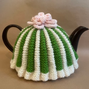 May include: A green and white striped knitted teapot cozy with a pink crocheted flower on top.