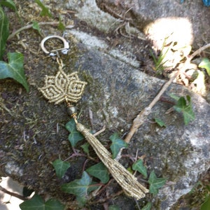 Könnte beinhalten: Goldfarbener Schlüsselanhänger mit einem Quasten und einem geknüpften Blumenanhänger. Der Anhänger ist aus goldfarbenem Kordel gefertigt und hat einen Perlenakzent.