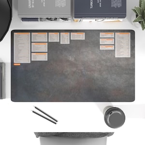 Peut inclure: Un tapis de bureau gris et orange avec une surface texturée. Le tapis présente une grille avec de petits carrés orange et du texte blanc. Le texte comprend les mots "Planification quotidienne", "Revue hebdomadaire", "Objectifs mensuels" et "Objectifs trimestriels".