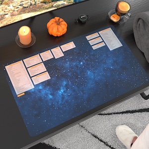 Puede incluir: Una alfombrilla de escritorio azul y negra con estampado de galaxia y cajas rectangulares naranjas con texto blanco. La alfombrilla está sobre una mesa negra con una calabaza, una vela y una taza de café.