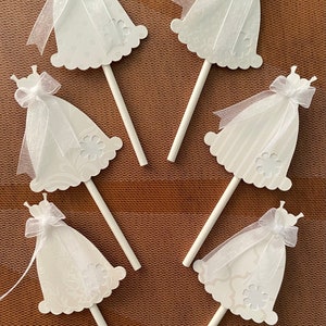 May include: Six white cupcake toppers shaped like wedding dresses with white tulle and bows. The toppers are on white sticks.