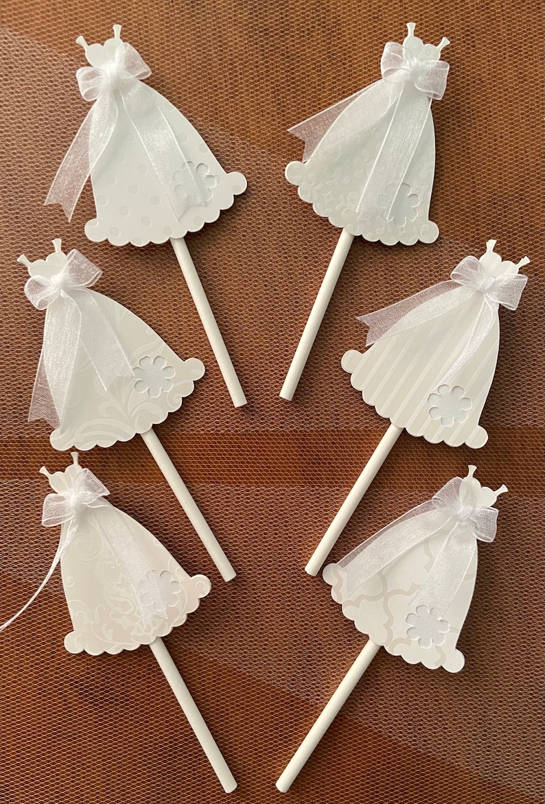 May include: Six white cupcake toppers shaped like wedding dresses with white tulle and bows. The toppers are on white sticks.