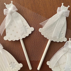 May include: Three white paper cupcake toppers shaped like wedding dresses with white ribbon bows and a white tulle overlay. The toppers are on white sticks.