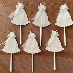 May include: Six white cupcake toppers shaped like wedding dresses with white ribbon bows and a white stick.