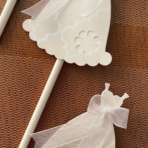 May include: Two white wedding bells with a white tulle bow and a white stick. The bells have a white floral pattern.